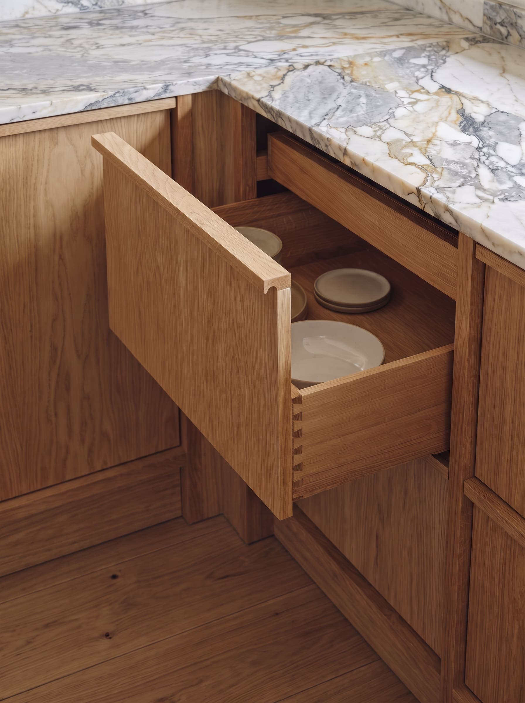 An open oak drawer under a marble worktop, with plates stacked inside.